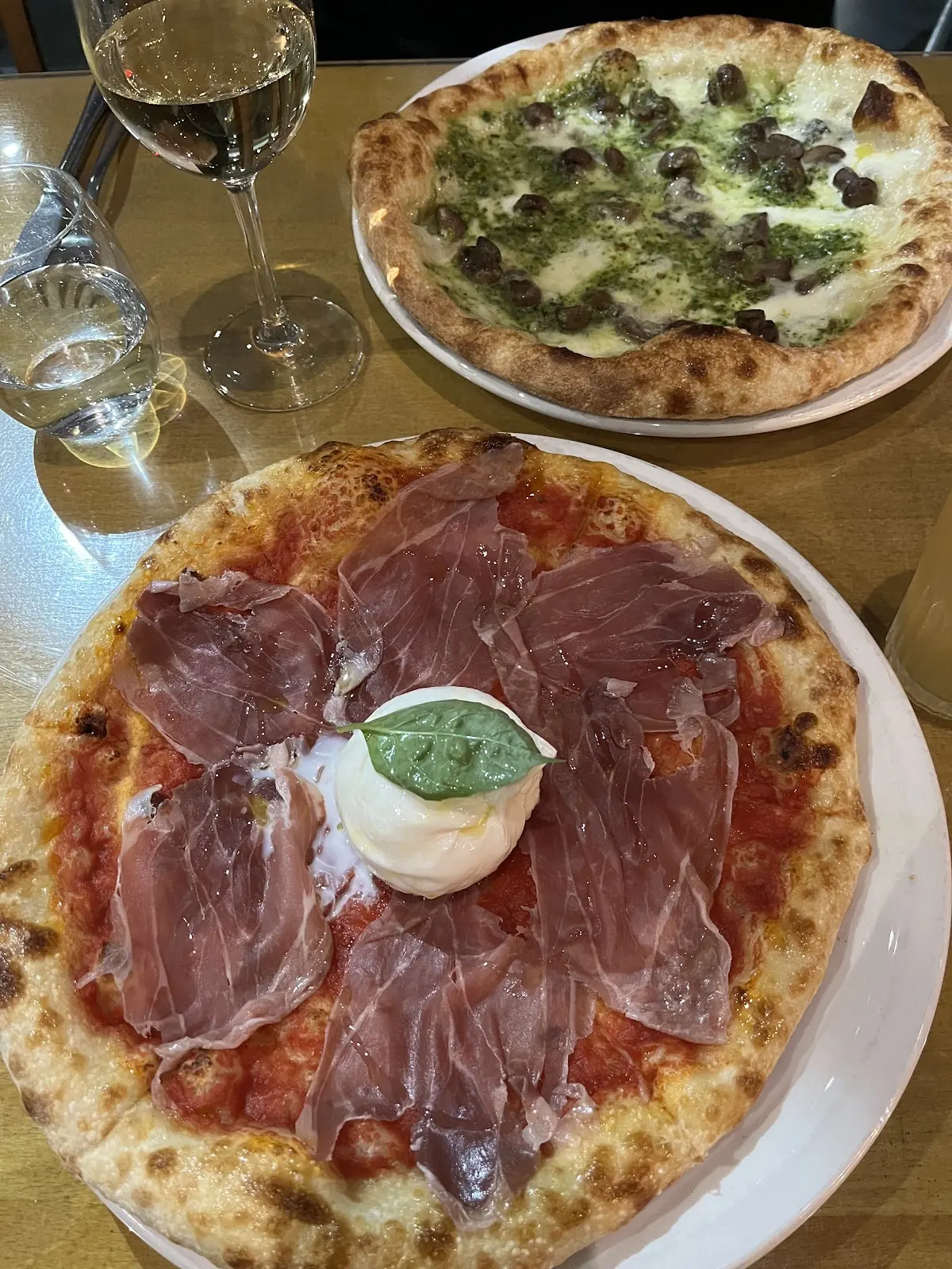 Two pizzas on the table, prosciutto and burrata in front, green pesto behind, with wine glasses
