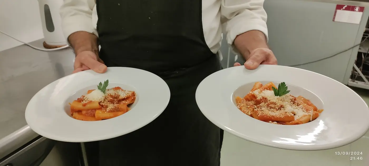A chef in whites carrying two plates of pasta in tomato sauce topped with basil, seen from above in the kitchen
